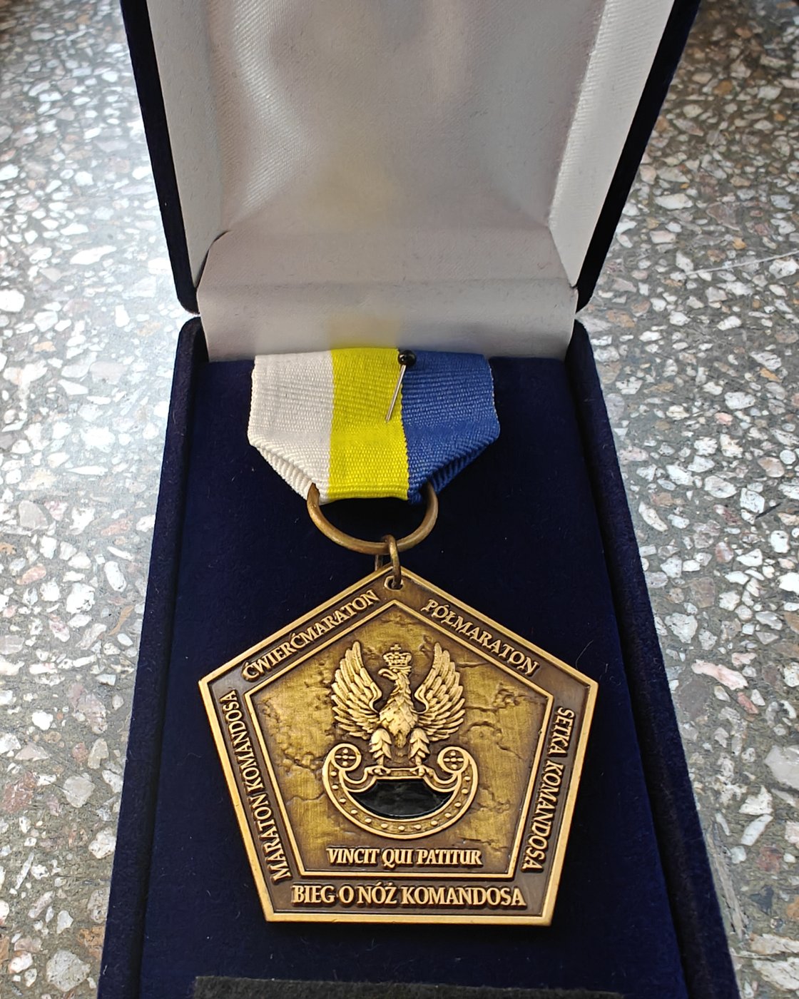 Medal Wielkiego Szlema Komandosa w granatowym etui: pięciokąt w kolorze antycznego złota z orłem wojsk specjalnych i napisem Vincit qui patitur, na biało-żółto-niebieskiej wstążce, poniżej baretka.