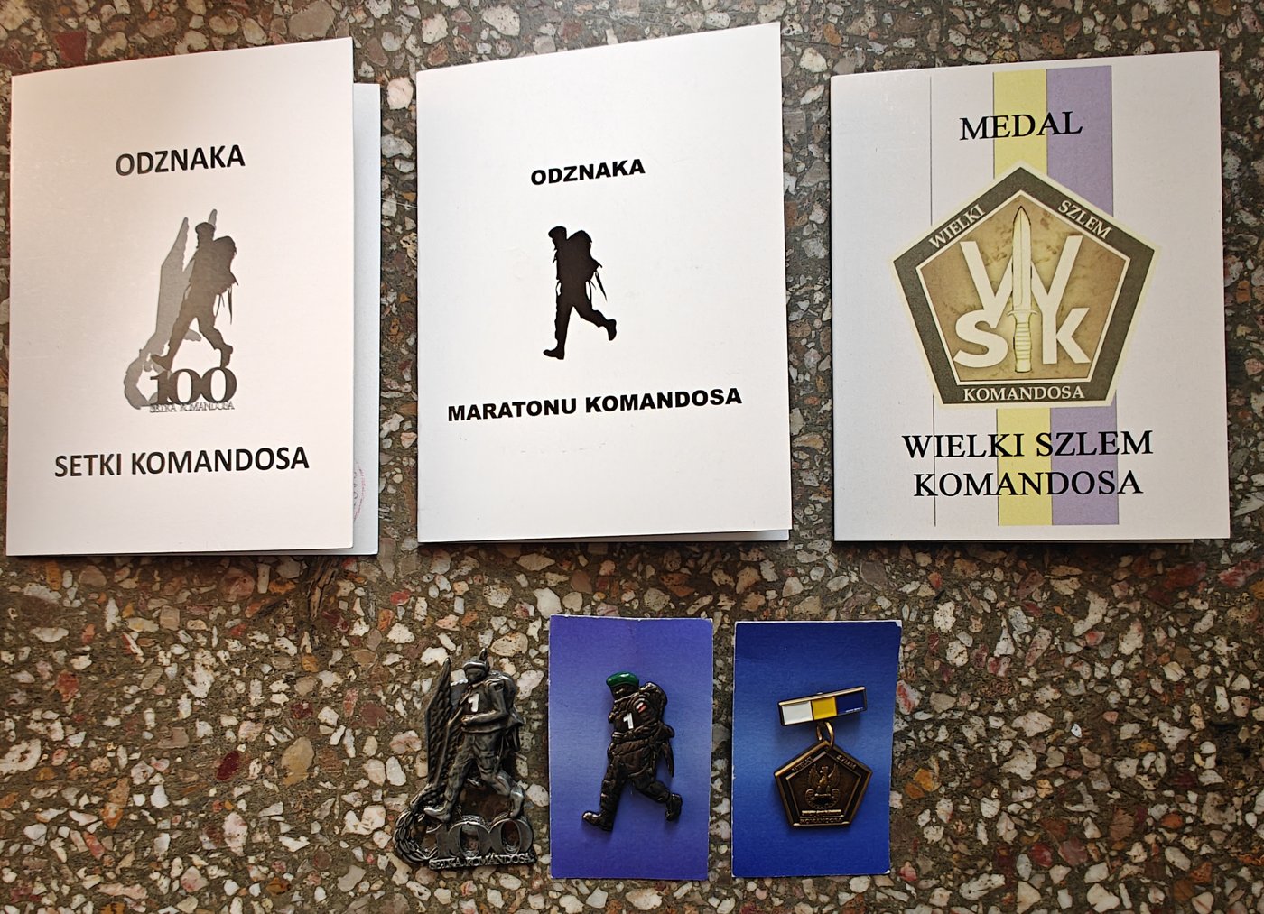 Legitymacje odznak Setki Komandosa i Maratonu Komandosa oraz medalu Wielkiego Szlema Komandosa, pod nimi obie odznaki i miniaturka medalu z baretką.